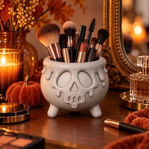 May include: A light gray ceramic cauldron-shaped holder featuring a raised skull design with dripping details and three small feet. The container is filled with various makeup brushes, including powder, blush, and spoolie brushes with metallic handles.
