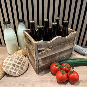Può includere: Una cassa di legno piena di bottiglie di vetro scuro con tappi dorati, accanto a due bottiglie di latte di vetro, pane, pomodori e un cetriolo. La cassa è su una superficie di legno con uno sfondo a strisce.