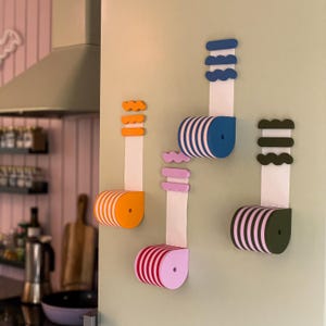 Könnte beinhalten: Vier farbenfrohe, an der Wand befestigte Papierhandtuchhalter. Jeder Halter hat eine einzigartige Farbe: Orange, Rosa, Blau und Grün. Die Halter haben ein zylindrisches Design mit weißen und farbigen Streifen und eine dekorative Oberseite. Das Papierhandtuch hängt herunter.
