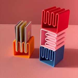 Op de afbeelding: Een oranje visitekaarthouder met een golvend ontwerp bevat meerdere kaarten. Rechts zijn drie kleurrijke, geometrische kaarthouders in rood, roze en blauw gestapeld. De houders hebben een uniek, harmonica-achtig ontwerp.