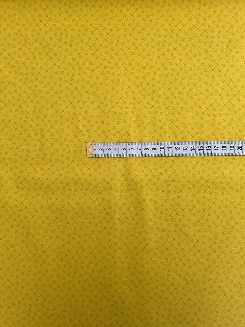 Puede incluir: Una tela amarilla con un patr&oacute;n repetitivo de peque&ntilde;as cruces marrones oscuras. Una regla se coloca sobre la tela, mostrando medidas en cent&iacute;metros.