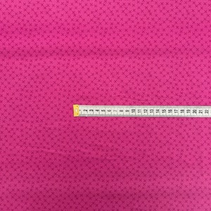 May include: A close-up of a pink fabric with a repeating pattern of small white crosses. A yellow measuring tape is placed across the fabric, showing measurements in inches.