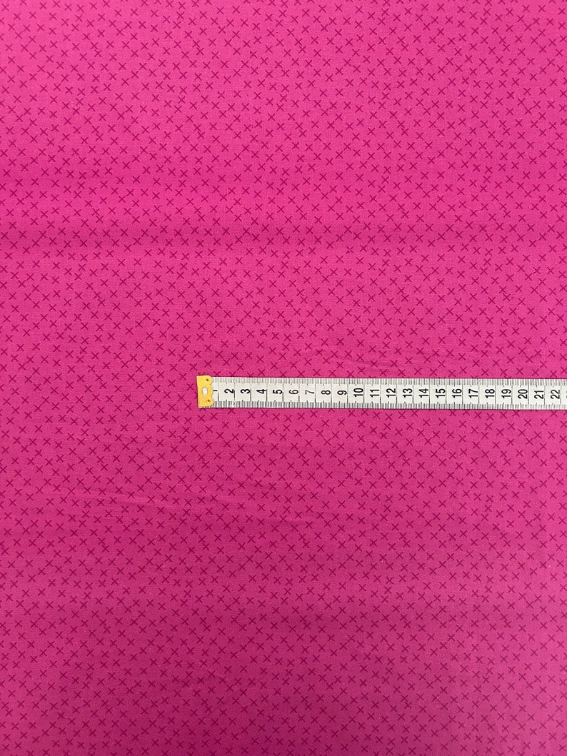 May include: A close-up of a pink fabric with a repeating pattern of small white crosses. A yellow measuring tape is placed across the fabric, showing measurements in inches.