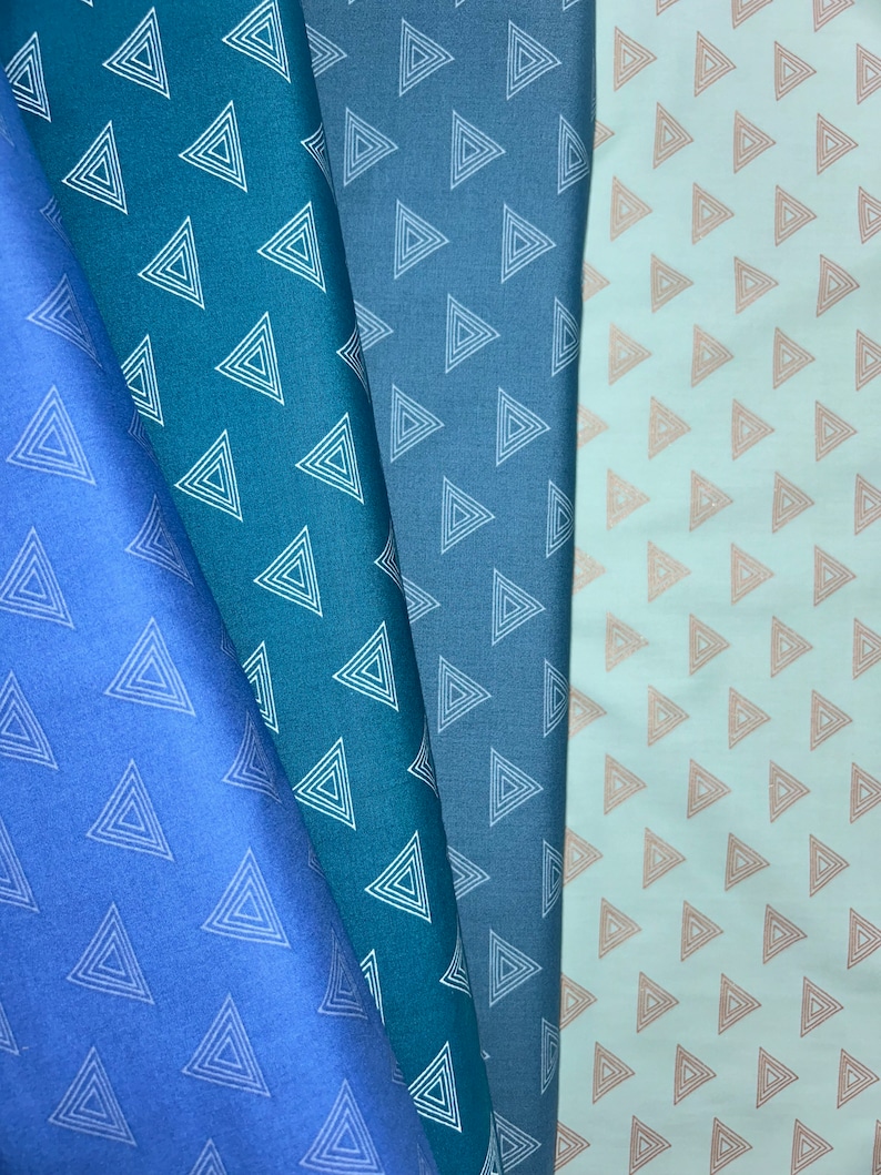 May include: Four fabric swatches in shades of blue and a light peach. Each swatch features a repeating pattern of white or peach-colored nested triangles. The fabrics are folded, showing the texture and color variations.