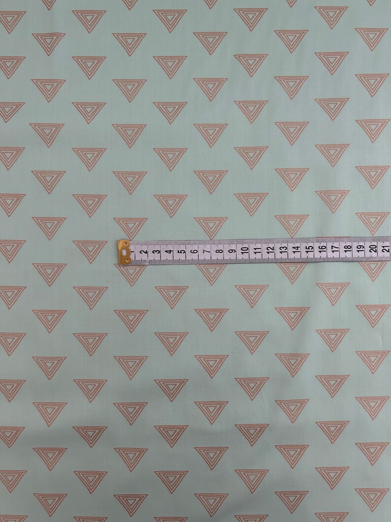 May include: Light blue fabric with a repeating pattern of nested, light brown triangles. A yellow and white measuring tape is laid across the fabric, showing measurements up to 21 inches.