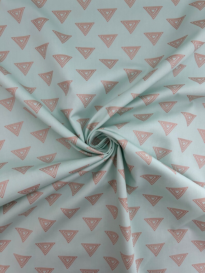 May include: A light blue fabric with a repeating pattern of brown outlined triangles. The triangles are arranged in a scattered layout across the fabric. The fabric is folded and draped, showing the texture and flow of the material.