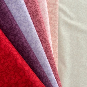 Könnte beinhalten: Eine Sammlung von sechs Stoffproben in verschiedenen Rosatönen und Lila mit einem kleinen Blumenmuster. Die Proben sind fächerförmig angeordnet, wobei der hellste Farbton oben und der dunkelste Farbton unten liegt.