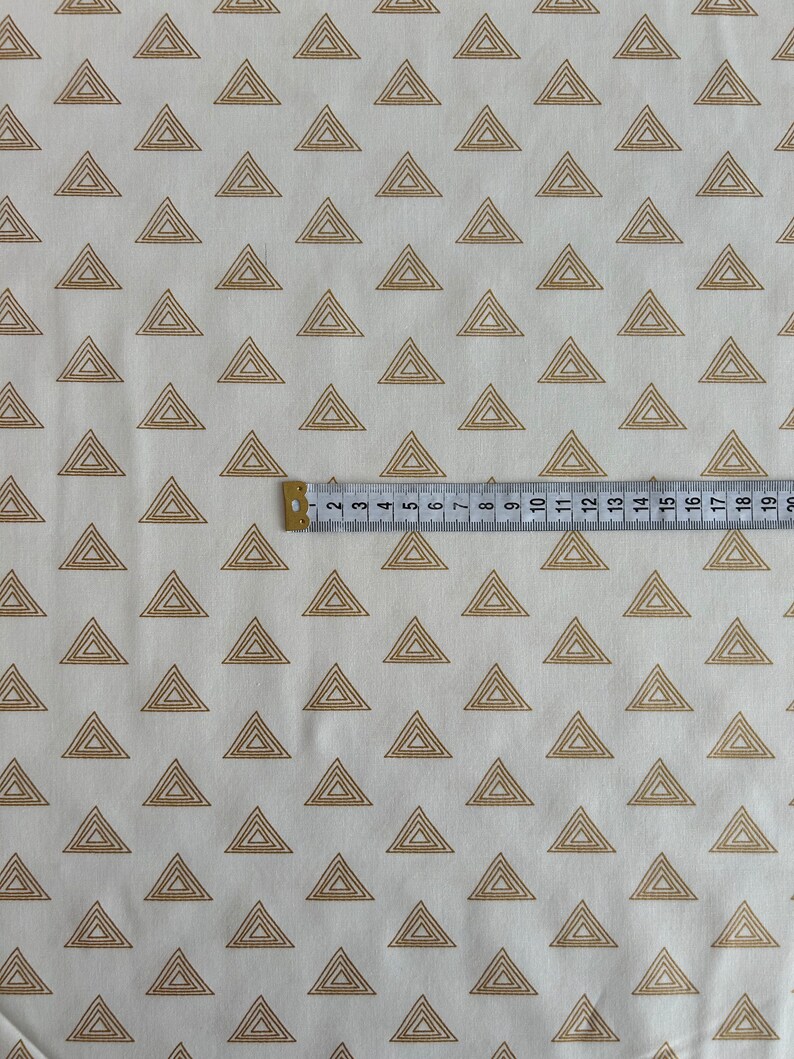 Puede incluir: Tela blanca con un patr&oacute;n repetido de tri&aacute;ngulos de contorno dorado. Los tri&aacute;ngulos est&aacute;n superpuestos y tienen un brillo sutil.