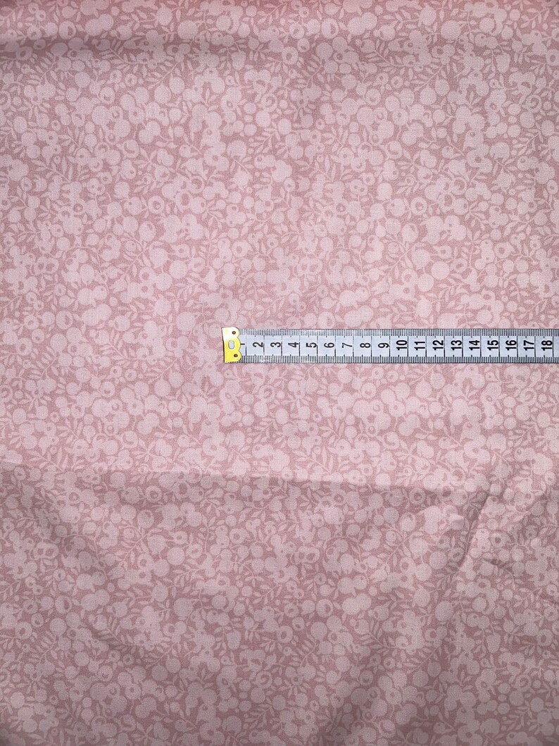 Może przedstawiać: Zbliżenie na r&oacute;żowy materiał z białym wzorem kwiatowym. Na materiale leży miarka, pokazująca wymiary w calach.