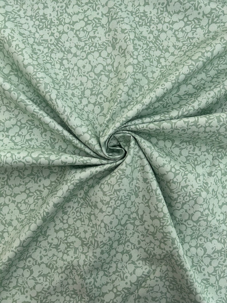 Pu&ograve; includere: Un tessuto verde salvia con un motivo floreale bianco. Il tessuto &egrave; piegato e ha una superficie morbida e testurizzata.