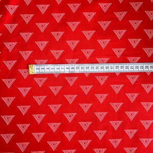 Może przedstawiać: Czerwona tkanina z powtarzającym się wzorem białych, zagnieżdżonych tr&oacute;jkąt&oacute;w. Taśma miernicza, oznaczona w centymetrach, jest ułożona na tkaninie. Odpowiednia do projekt&oacute;w krawieckich lub rękodzielniczych.