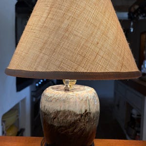 May include: A table lamp featuring a conical, textured tan burlap lampshade with a dark brown trim. The base is a rounded ceramic vessel with a reactive glaze finish, transitioning from a dark brown bottom to a white and light brown top section.