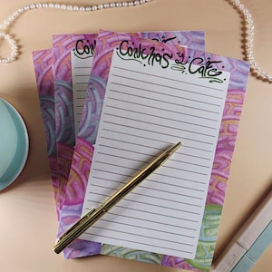 May include: A stack of rectangular lined paper notepads featuring a colorful pattern of pink, purple, and green circular shapes. The top sheet displays the text Conchas y Café in black script at the top, with a gold ballpoint pen resting on it.