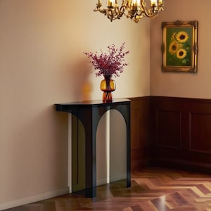 Puede incluir: Una consola negra moderna con patas arqueadas sostiene un jarrón de bayas rojas. El jarrón de color ámbar está debajo de una lámpara de araña dorada y una pintura de girasoles enmarcada. La mesa está contra una pared beige.