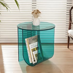 May include: A modern, teal-colored side table with a circular top and a built-in magazine rack. The table holds several books and a small white vase with dried flowers. The table is made of a transparent material.