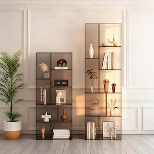 Puede incluir: Dos estanterías de exhibición de metal negro y vidrio con varios objetos decorativos. Las estanterías contienen libros, jarrones, esculturas y un reloj que muestra las 12:59. Una planta en maceta es visible a la izquierda.