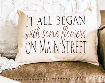 Puede incluir: Una almohada blanca con el texto "IT ALL BEGAN with some flowers ON MAIN STREET" en letras negras. El texto "with some flowers" está en color marrón.