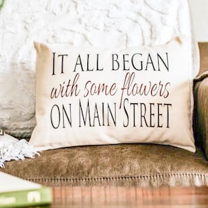 Cotton love story pillow sitting on brown leather couch, pillow reads it all began with some flowers on Main Street. Pillow is customizable to your love story. Sized 12x16 inches. Available on multiple sizes and fabrics.