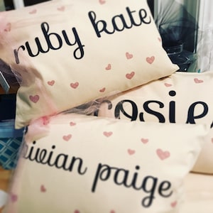 May include: Three white pillowcases with pink hearts printed on them. Each pillowcase has a name written in black lettering: "ruby kate", "cosie", and "iviian paige".