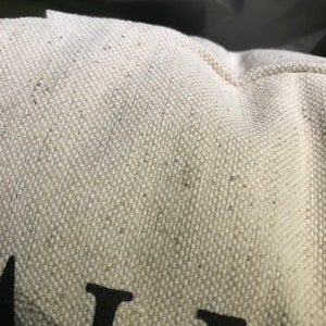 May include: Close-up of a white canvas tote bag with black text that reads "ALL". The bag has a subtle texture and is made of a natural fabric.