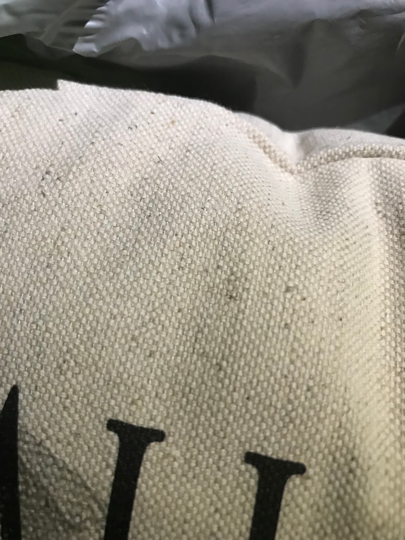 May include: Close-up of a white canvas tote bag with black text that reads "ALL". The bag has a subtle texture and is made of a natural fabric.