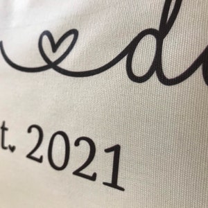 May include: White canvas pillow with black text that reads "Mom & Dad Est. 2021" with a heart symbol between the words "Mom" and "Dad".
