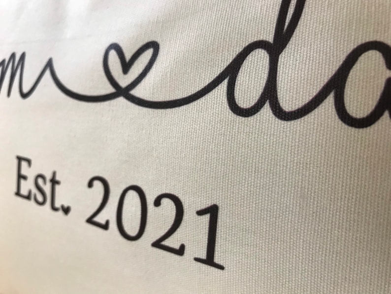 May include: White canvas pillow with black text that reads "Mom & Dad Est. 2021" with a heart symbol between the words "Mom" and "Dad".