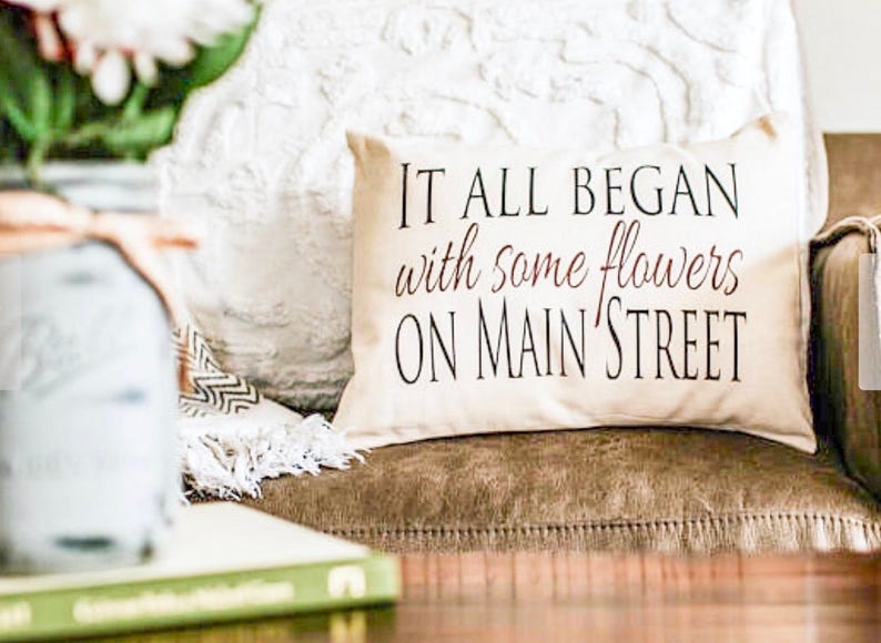 Cotton love story pillow sitting on brown leather couch, pillow reads it all began with some flowers on Main Street. Pillow is customizable to your love story. Sized 12x16 inches. Available on multiple sizes and fabrics.