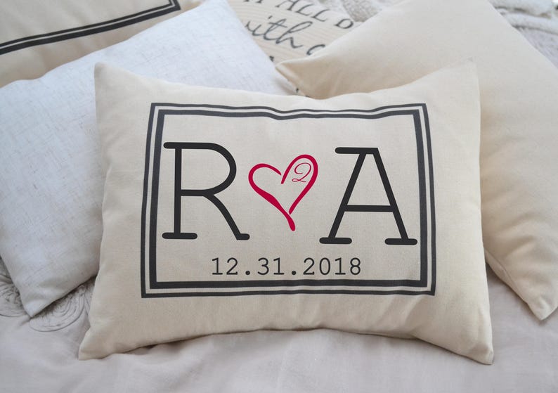 Cotton decor cream colored pillow sized at 12x16 inches. Pillow has black frame, with two large initials R & A separated by a heart with the number 2 inside the top right curve of the heart. 8 digit date at the bottom separated by periods.