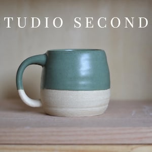 May include: A ceramic mug with a rounded shape featuring a sage green glazed upper section and a matte beige lower section. The text STUDIO SECONDS is printed in white capital letters above the mug. The mug has a curved handle on the left side.