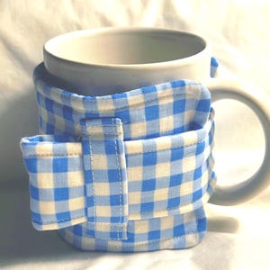 May include: A white ceramic mug wrapped in a light blue and white gingham fabric sleeve. The sleeve features a horizontal band with a vertical tab closure, secured with stitching along the edges, providing a snug fit around the mug's exterior.