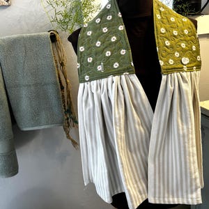May include: A hanging kitchen towel featuring a quilted olive green top panel with a white daisy print and a white button closure. The lower section consists of a gathered, light beige and white vertical striped fabric that hangs in soft folds.