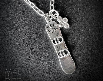Può includere: Una collana d'argento con un ciondolo a forma di snowboard e un design a fiocco di neve. Il ciondolo ha alcuni piccoli cristalli trasparenti attaccati.