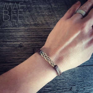Puede incluir: Un brazalete de cuero marr&oacute;n con un acento de metal trenzado. El brazalete se lleva en la mu&ntilde;eca.