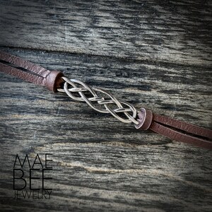 Puede incluir: Un brazalete trenzado plateado con correas de cuero marr&oacute;n. El brazalete est&aacute; sobre una superficie de madera.