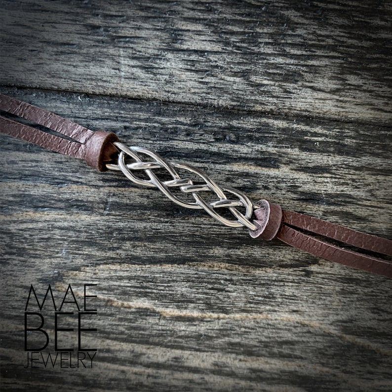 Puede incluir: Un brazalete trenzado plateado con correas de cuero marr&oacute;n. El brazalete est&aacute; sobre una superficie de madera.