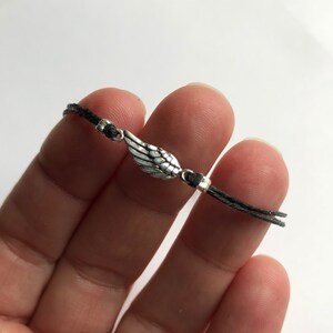 以下が含まれることがあります： 銀色の羽のチャームが付いた繊細なブレスレット。チャームは黒いコードの中央に配置され、小さな銀色のビーズでコードが固定されています。ブレスレットはミニマリストなデザインです。