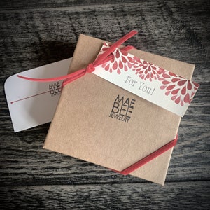 Puede incluir: Una caja de regalo marr&oacute;n con una cinta roja y una etiqueta de regalo que dice "&iexcl;Para ti!" La etiqueta tiene un dise&ntilde;o floral rojo y blanco. La caja est&aacute; etiquetada con "Mae Bee Jewelry".