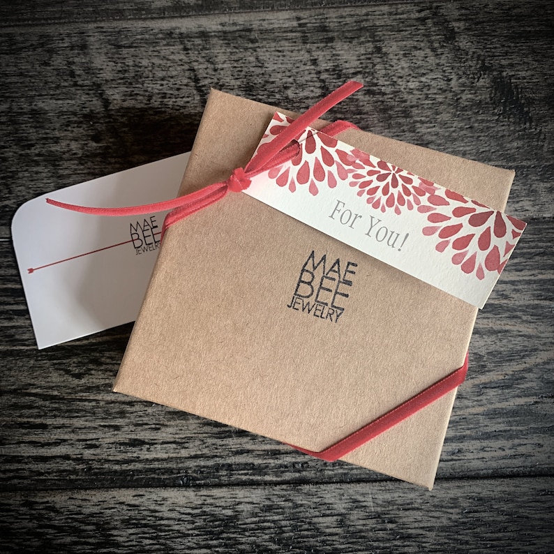 Puede incluir: Una caja de regalo marr&oacute;n con una cinta roja y una etiqueta de regalo que dice "&iexcl;Para ti!" La etiqueta tiene un dise&ntilde;o floral rojo y blanco. La caja est&aacute; etiquetada con "Mae Bee Jewelry".