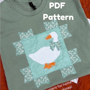 May include: A sage green t-shirt featuring a central square appliqué of a white goose with an orange beak and feet, wearing a floral bow. The design includes the text PDF Pattern and Excellent for home sewing machines! in white lettering.