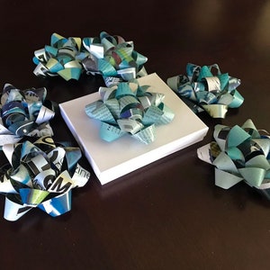May include: Six blue and white paper gift bows with a white box in the center. The bows are made from recycled magazine pages.