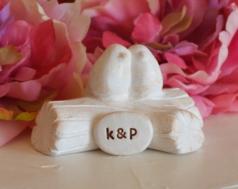 May include: White ceramic wedding cake topper featuring two birds perched on a log with the initials "K & P" engraved on the log. The cake topper is surrounded by pink and purple flowers.