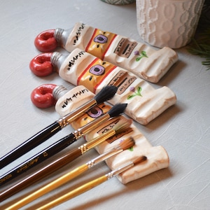 May include: A ceramic paint brush rest shaped like three squeezed paint tubes with red caps, featuring hand-painted details in yellow, purple, and green. Five artist paint brushes with black, brown, and gold handles rest across the tubes.
