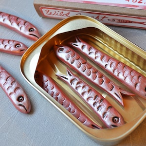 May include: A close-up of a tin of sardines with several fish-shaped objects. The fish are reddish-brown with detailed scales and fins. The tin is gold-colored, and a cardboard box with the text "Wild Delight" is visible.