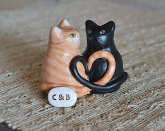 May include: Ceramic figurine of two cats, one orange and white and one black, with their tails intertwined to form a heart shape. The base of the figurine is white with the text "C&B" written on it.