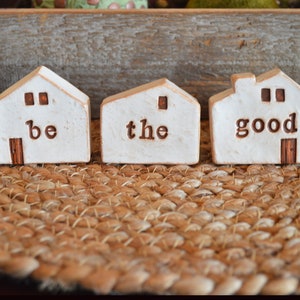 May include: Three small white ceramic houses with brown accents. The houses are arranged in a row and each has a word inscribed on the front: "be", "the", and "good".