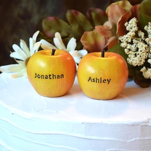 May include: Two yellow and orange decorative apples with brown stems, each featuring a black printed name. One apple is labeled Jonathan and the other is labeled Ashley. The apples are positioned on a white surface with white flowers.