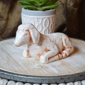May include: A cream-colored ceramic lamb figurine in a resting pose. The lamb has detailed features and reddish-brown accents. It sits on a round, distressed wooden tray with a metal rim. A small potted succulent is in the background.