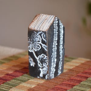May include: A decorative wooden block with a house-like shape. The block is painted black with white floral designs on one side and vertical white lines on another. The top is a light brown color with horizontal lines.