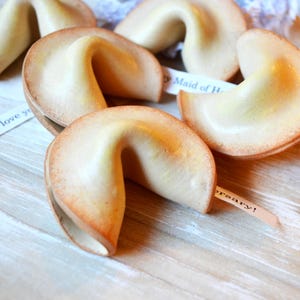 May include: Several fortune cookies with light brown edges and a pale yellow interior are arranged on a light wood surface. Some cookies have small paper strips with text peeking out. The text on one strip reads "Anniversary!"